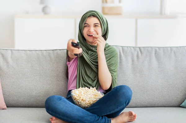 Divertimenti a domicilio. Piena lunghezza di musulmana adolescente in hijab con telecomando mangiare popcorn e guardare la TV — Foto Stock