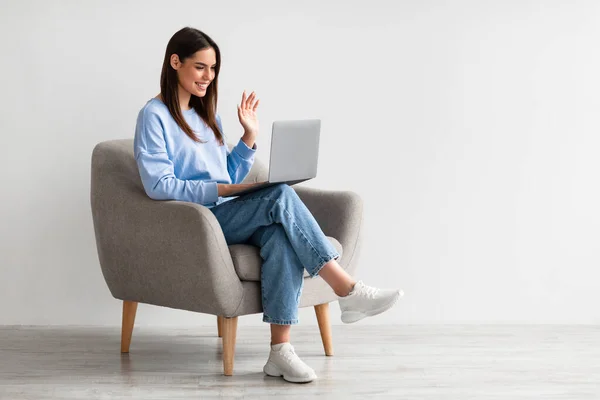 Retrato de comprimento total da senhora caucasiana usando laptop para comunicação on-line em poltrona contra a parede do estúdio branco — Fotografia de Stock