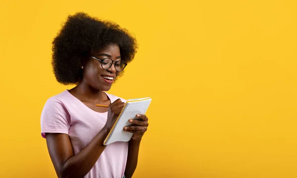 Smart african american female student in glasses writing checklist, taking notes in copybook over yellow background — Stock Photo, Image