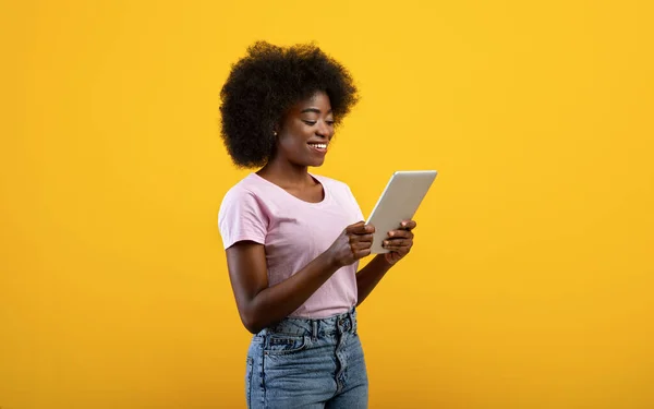 Young black lady with modern digital tablet, using mobile application on pad, posing on yellow studio background — Stock Photo, Image