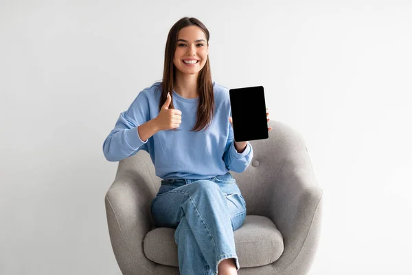 Γοητευτική χιλιετή γυναίκα δείχνει tablet PC με άδεια οθόνη, gesturing αντίχειρα επάνω στην πολυθρόνα, mockup για το σχεδιασμό — Φωτογραφία Αρχείου