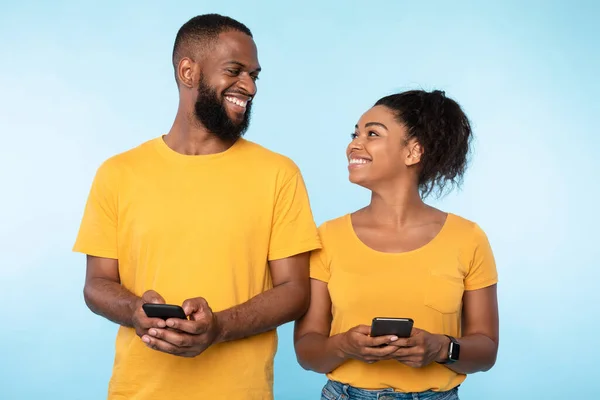 Comunicação de pessoas milenares. Mulher negra e homem mensagens de texto em telefones celulares, de pé lado a lado no fundo azul — Fotografia de Stock