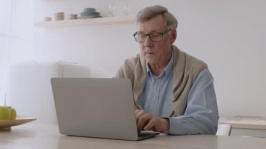 Dizüstü bilgisayarda yazan, gözlüğünü çıkaran ve sinir bozucu bir şekilde bilgisayara bakan kıdemli bir adam.