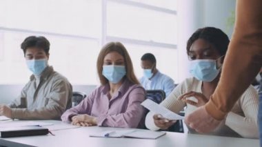 Öğretmen koruyucu tıp maskeli öğrencilere testler yapıyor, grup arkadaşları sınıfta birbirlerine çarşaf dağıtıyor.