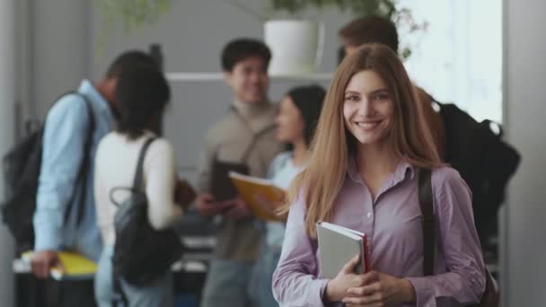 Bonito adolescente caucasiano menina sorrindo para a câmera, posando com livro e mochila contra seus diversos colegas de grupo, espaço vazio — Vídeo de Stock