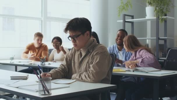 Trauriger unterdrückter asiatischer Kerl Nerd sitzt abseits von Klassenkameraden, Studenten werfen Papierbälle auf ihn und lachen — Stockvideo