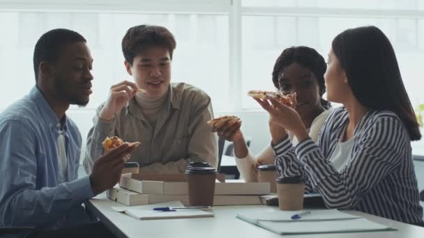 Gruppo di studenti multietnici positivi che mangiano pizza consegnata e discutono di educazione, ridendo insieme a tavola — Video Stock