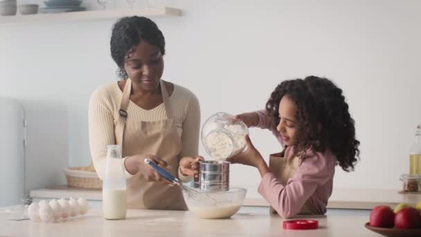 Familie bakkerij. Klein Afrikaans amerikaans meisje helpen haar moeder met het bereiden van deeg, het toevoegen van bloem te zeven in de keuken — Stockvideo