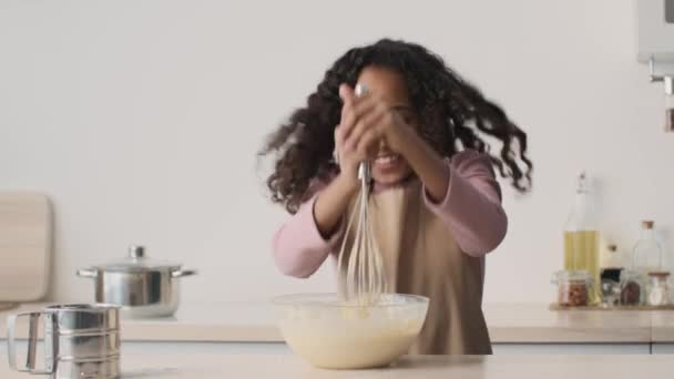 장난 스러운 작은 아프리카 계 미국인 여자 애 앞치마를 입고 반죽을 반죽에 넣고 위스키를 그릇에 넣고 점프하고 웃고 — 비디오