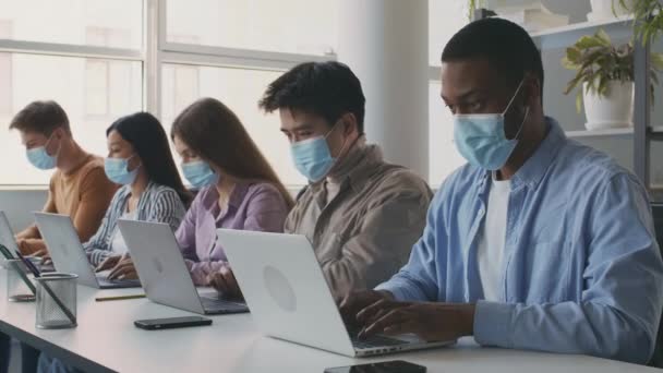 Groupe de jeunes étudiants multiethniques portant des masques médicaux assis en classe et tapant rapidement sur leur ordinateur portable — Video