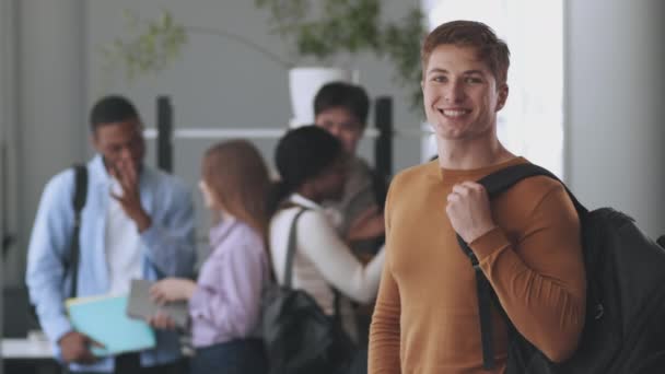 Cara confiante com mochila sorrindo para a câmera, posando na faculdade sobre conversar com vários colegas de grupo no fundo — Vídeo de Stock