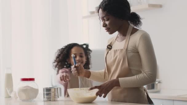 Jovem senhora afro-americana preparando massa na cozinha, sua filha chegando e degustando pastelaria crua, abraçando e rindo — Vídeo de Stock