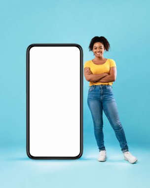 Güzel Afro kadın, mavi arka planda beyaz ekranı boş, kocaman bir akıllı telefonun yanında duruyor. En yeni mobil uygulama