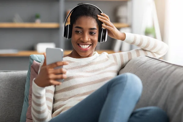 Młoda czarna kobieta w słuchawkach bezprzewodowych Słuchanie muzyki na smartfonie w domu — Zdjęcie stockowe