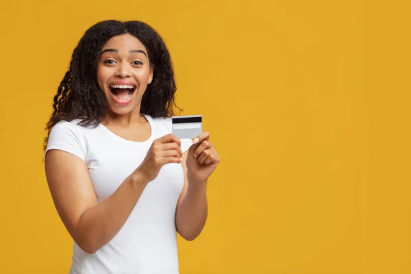 Extasiado Africano americano senhora segurando cartão de crédito na mão, posando sobre fundo estúdio amarelo com espaço de cópia — Fotografia de Stock