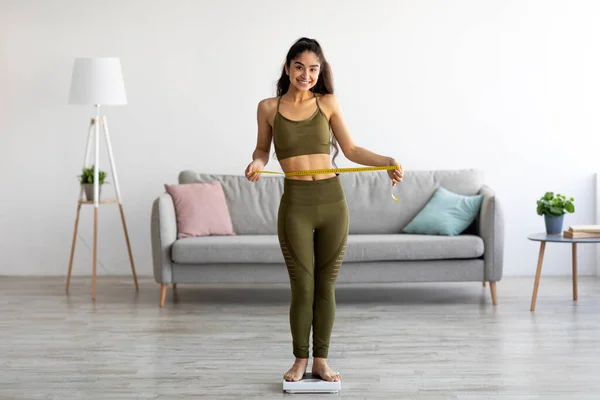 Asiática dama en ropa deportiva de pie en balanzas, la medición de la cintura con cinta, sentirse feliz por perder peso en casa — Foto de Stock