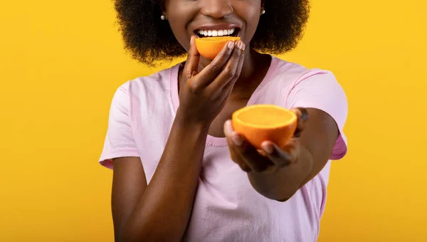 Sommar detox koncept. Afroamerikansk kvinna bita orange hälften och sträcker andra till kameran, gul bakgrund — Stockfoto