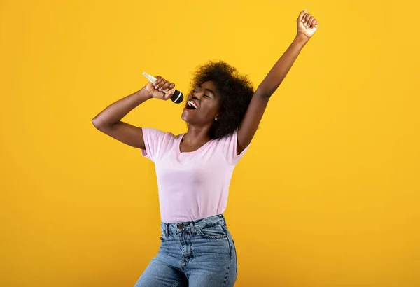 Müzik aşığı. Heyecanlı siyah kadın mikrofonda şarkı söylüyor ve dans ediyor, sarı arka planda eğleniyor. — Stok fotoğraf