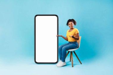 Mobil uygulama veya web sitesi tasarımı için modeli olan boş akıllı telefonun yanında oturan Afro kadın, mavi arkaplan