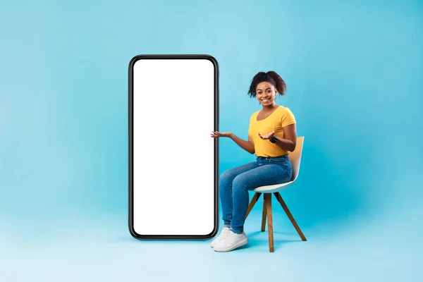 Mobil uygulama veya web sitesi tasarımı için modeli olan boş akıllı telefonun yanında oturan Afro kadın, mavi arkaplan — Stok fotoğraf