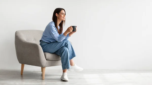 Full längd av ung kvinna dricker varmt kaffe i mysig fåtölj mot vit studio vägg, banner med fritt utrymme — Stockfoto