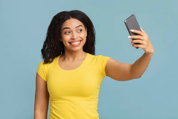 Pozitif siyah kadın akıllı telefondan özçekim yapıyor, poz veriyor ve mavi stüdyo arka planında eğleniyor. — Stok fotoğraf