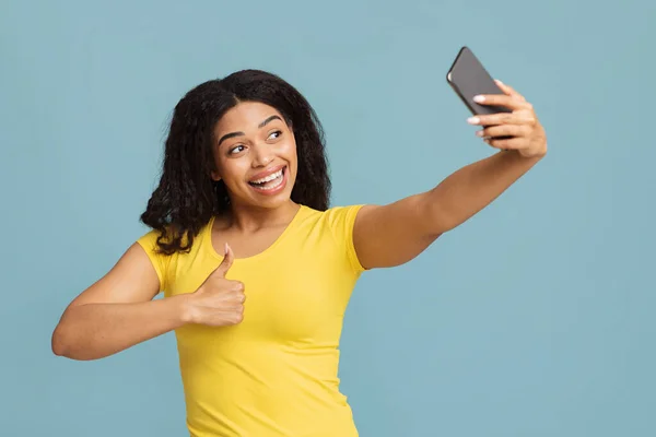 Heyecanlı Afrikalı Amerikalı kadın akıllı telefondan selfie çekiyor ve mavi arka planda baş parmağını gösteriyor. — Stok fotoğraf