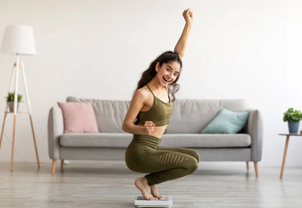 Donna indiana trionfante in abiti sportivi seduta su bilance, gesticolando SI, soddisfatta del suo risultato di perdita di peso — Foto Stock