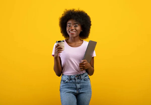Happy black female student with laptop computer and takeaway coffee posing to camera on yellow background — Stock Photo, Image