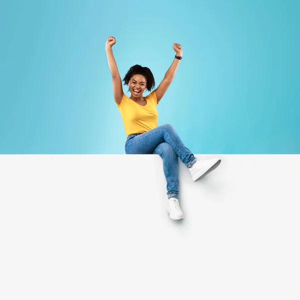 Portrait of excited young black lady sitting on blank white paper banner with mockup, raising hands up