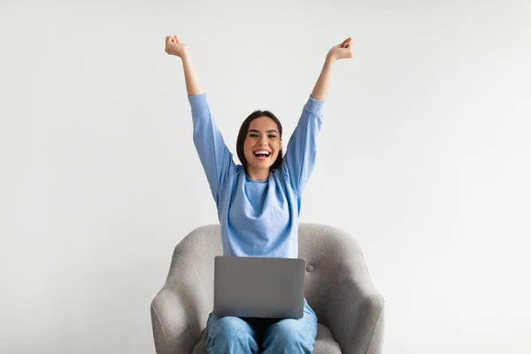 Ευφορική νεαρή γυναίκα γιορτάζει την επιτυχία με φορητό υπολογιστή, σηκώνοντας τα χέρια, κάθεται στην πολυθρόνα σε λευκό φόντο — Φωτογραφία Αρχείου
