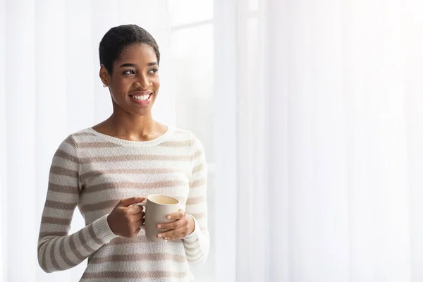 Gelukkig glimlachende Afro-Amerikaanse vrouw drinken koffie terwijl staande in de buurt van venster thuis — Stockfoto