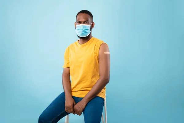 Vaccinated black man in face mask wearing plaster bandage on shoulder after covid-19 vaccine injection, blue background