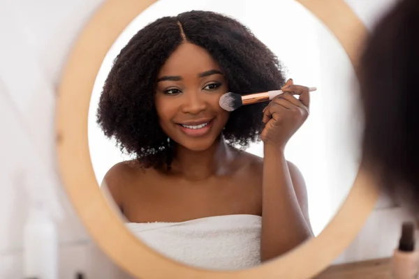 Bonita dama negra aplicando maquillaje cerca del espejo, usando cosméticos decorativos en interiores —  Fotos de Stock