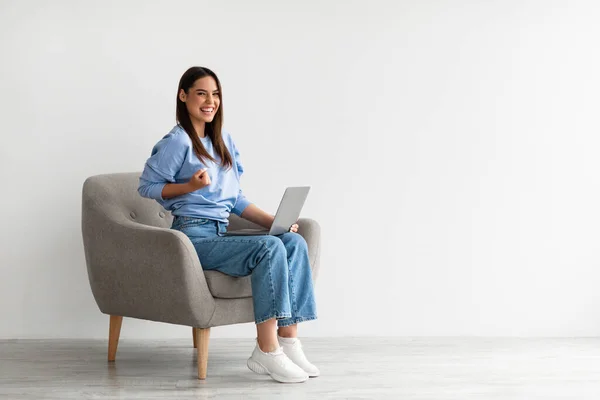 Glada unga kvinna sitter i fåtölj med bärbar dator, gör JA gest, få goda nyheter på nätet, uppnå framgång — Stockfoto