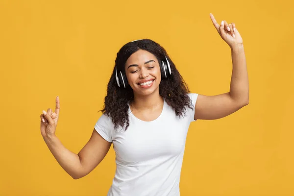 Bouge ton corps. Femme afro-américaine positive écoutant de la musique dans des écouteurs sans fil et dansant, fond jaune — Photo