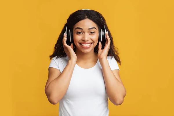 Femme afro-américaine excitée dans un casque sans fil écoutant de la musique, profitant d'un bon son sur fond jaune — Photo