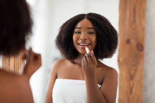 Potret wanita muda Afro yang mempesona mengaplikasikan lipstik dan melihat ke cermin di rumah. Konsep makeup organik — Stok Foto