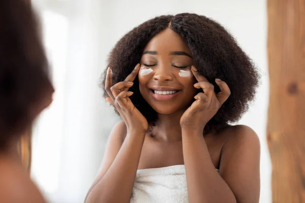 Encantadora afro dama milenaria aplicando crema facial nutritiva bajo sus ojos cerca del espejo en casa —  Fotos de Stock