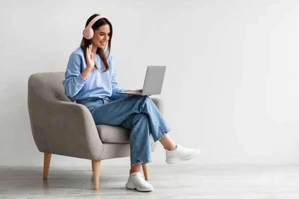 Mujer alegre en auriculares usando el ordenador portátil, teniendo conferencia en línea, sentado en sillón, saludando a la webcam —  Fotos de Stock