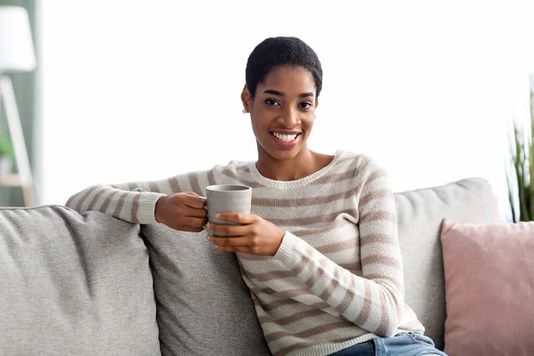 Εβδομαδιαίο διάλειμμα. Χαρούμενη μαύρη κυρία με φλιτζάνι καφέ χαλαρώνοντας στον καναπέ — Φωτογραφία Αρχείου
