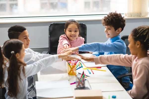 Happy multiethnic schoolchildren putting hands together at classroom — Stock Photo, Image