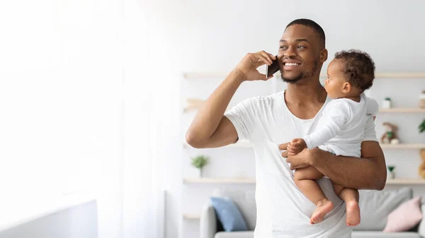 Negro hombre celebración pequeño bebé en las manos y hablando en celular —  Fotos de Stock