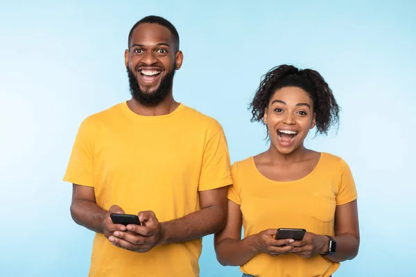 Vida online. Millennial casal afro usando telefones celulares, verificando web ou mídias sociais em fundo estúdio azul — Fotografia de Stock