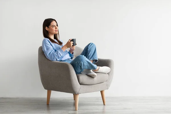 Jovem positiva sentada em poltrona com pernas cruzadas, aquecendo as mãos na xícara de café quente, desfrutando de bebida quente — Fotografia de Stock