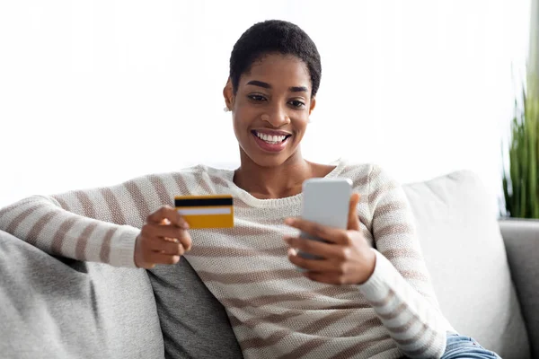 Aplicación bancaria. Joven dama negra con Smartphone y tarjeta de crédito en casa —  Fotos de Stock