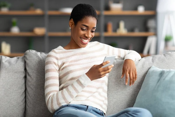 Sorrindo mulher negra navegando novo aplicativo no smartphone enquanto descansa em casa — Fotografia de Stock