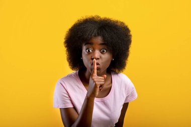 Sessiz olun. Siyah kadın kameraya sessiz jest yapıyor, sarı arka planda duruyor, stüdyo çekimi.