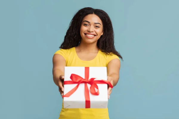 问候和庆祝。快乐的黑人女士拿着包装好的礼品盒，把礼物伸向相机，蓝色背景 — 图库照片