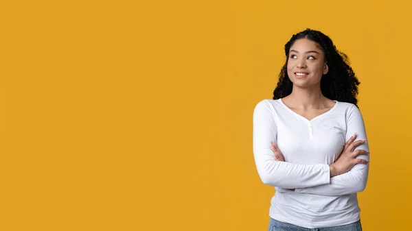 Мечтательная молодая афроамериканка, стоящая со сложенными руками на жёлтом фоне — стоковое фото